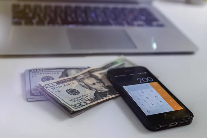 A phone with a calculator app open sits on a desk with cash and a personal computer