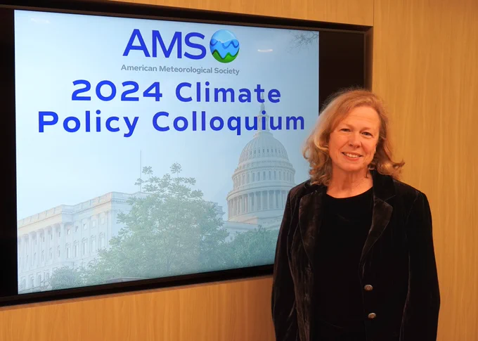 A woman stands in front of a presentation screen displaying the logo of the American Meteorological Society and text reading "2024 Climate Policy Colloquium" with an image of the U.S. Capitol building.