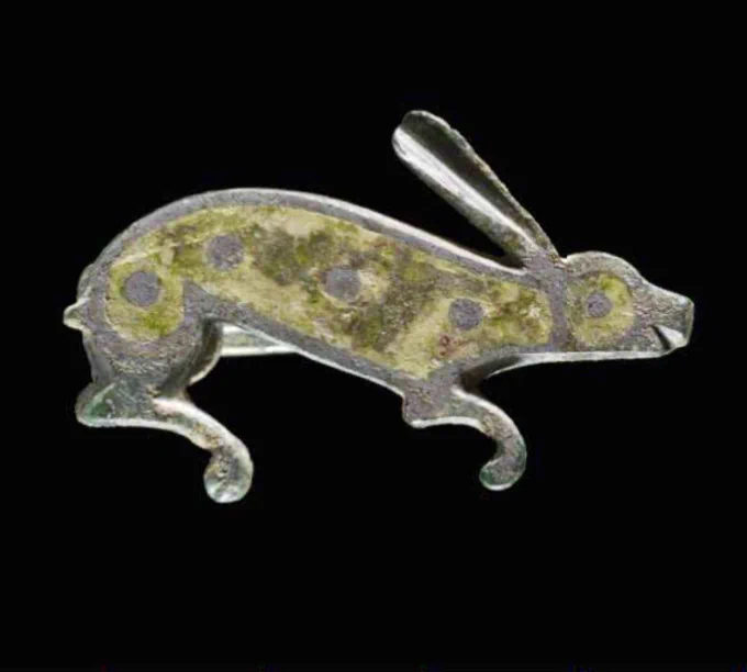Romano-British brooch in the form of a crouching hare, viewed from the side with head to the right. The hare has one long ear, a slightly curved incised mouth, and two curved legs (one back and one front). The brooch is cast from copper alloy (bronze), now grey in colour. The body is inlaid with green enamel with four copper-alloy decorative dots in the green enamel. The head is decorated with a circular green enamel inlay, at the centre of which is a copper-alloy dot for the eye.