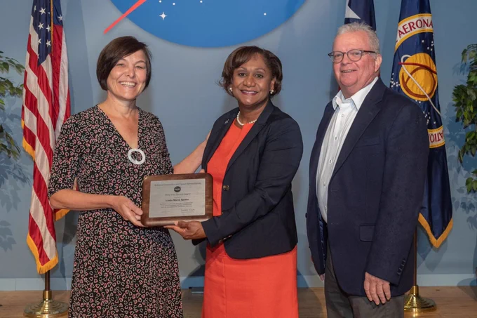Linda Spuler, part Ojibwe, smiles at the camera as she receives her 30-year service award from NASA Johnson Director Vanessa Wyche. Johnson Center Operations Director Joel Walker stands to Wyche’s right. A United States flag and a NASA flag are behind the group. The bottom portion of NASA’s meatball insignia can be seen on the wall behind them. Credit: NASA/David DeHoyos