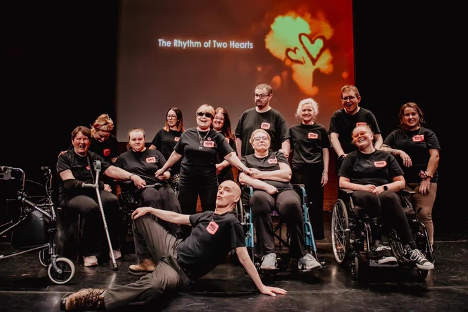 People on a stage in Black T-Shirts. Some in chairs some standing and one reclined on the floor.