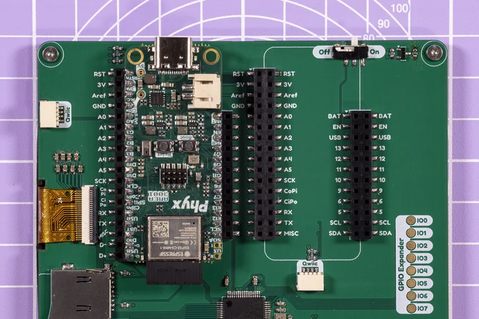 A photo of a pcb on a lavender cutting matt. The PCB has two slots for Feather boards. One slot is taken by the Tess board. There is also a flex cable sneaking in from the other side of the board and into a fpc connector. There is an on/off switch. There's a part of a microSD connector, there are two Qwiic connectors, one facing outwards and one facing inwards. There's also a part of a QFP chip visible. And lastly there are 8 test points marked with IO0-IO7 and "GPIO Expander" next to them.