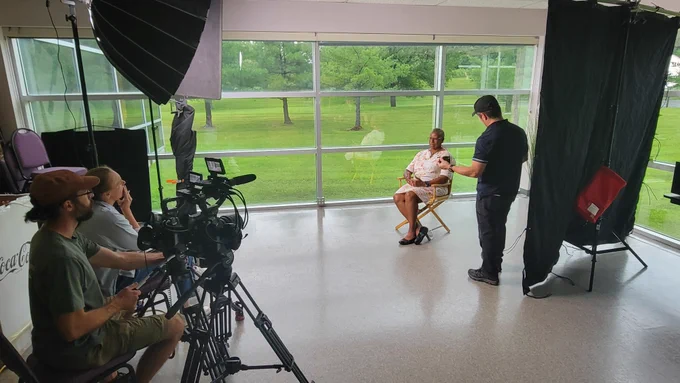 A woman sits in a director's chair waiting to be interviewed for a TV production. Two videographers and their equipment are visible in the foreground. A production assistant stands near her chair. She is sitting in a room with a wall of windows. 