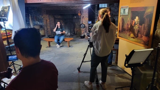 A woman wearing a Parks polo shirt sits on a bench in the kitchen of 1700s home Crailo. Behind her is a large open-hearth brick fireplace with implements typical of the period. Two videographers are in the foreground arranging the shot. The woman has her hand next to her ear as if she is adjusting an earpiece. 