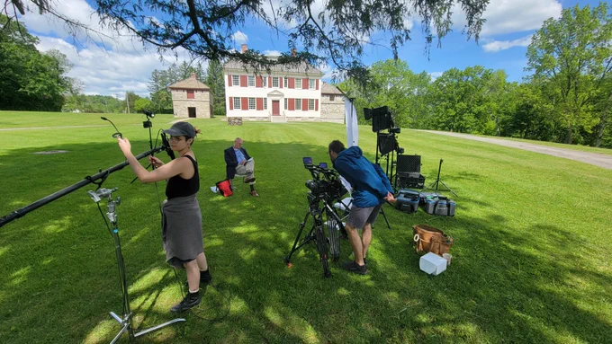 A man sits on a chair on the lawn of Johnson Hall State Historic Site while two videographers arrange equipment. It is a summer day and the two-story colonial-style home is visible behind him. 