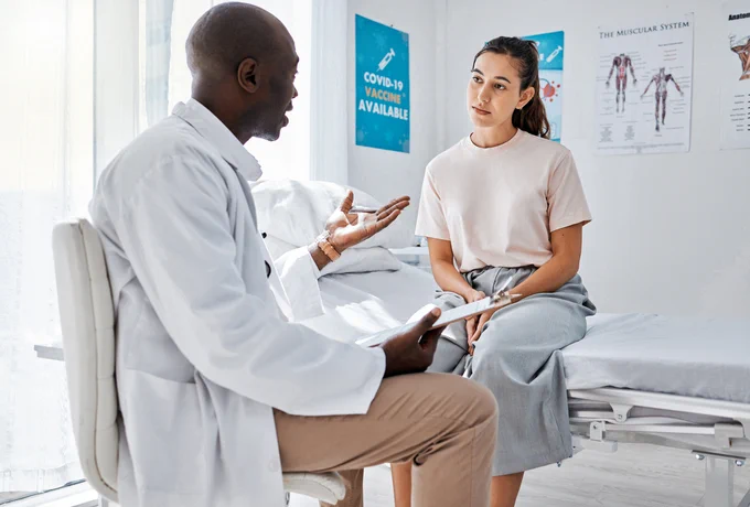 A healthcare professional in a white coat is discussing with a patient who is seated on an exam table in a medical office with posters including one titled "COVID-19 Vaccine Available.