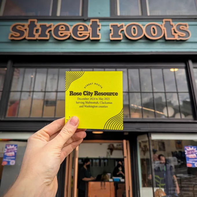 A hand holds up a square, yellow booklet with black text on the cover. The text says, "Street Roots Rose City Resource. December 2024 to May 2024. Serving Multnomah, Clackamas, and Washington Counties. Behind the booklet is the none "Street Roots" sign on the front of the Street Roots office building.