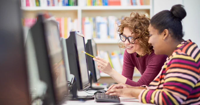 Two individuals collaborating at a computer in a library setting.