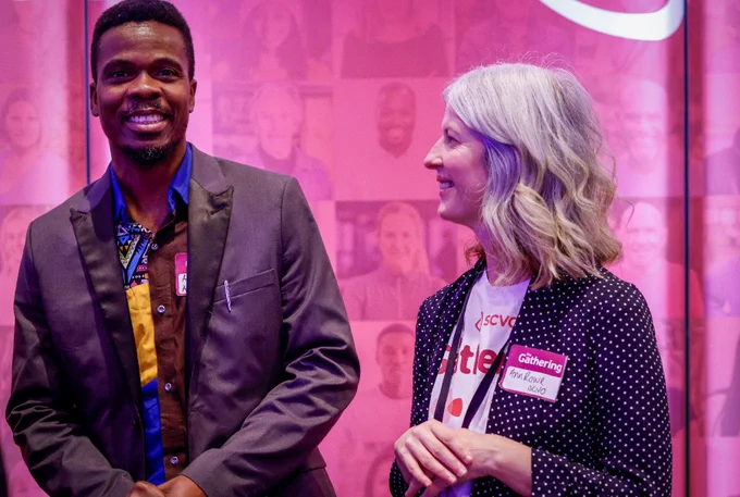 A man smiles and faces the camera. He wears a suit jacket and colourful shirt. A woman with curly hair, in a polka dot blazer and a white t-shirt smiles at the man. Both people wear a name badge with the event title "the Gathering". They are in front of a banner with photos depicting SCVO members
