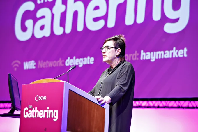 Anna Fowlie, dressed in black top, with short dark hair and wearing glasses stands at a lectern with a graphic that reads "SCVO the Gathering". She's addressing an audience. Anna stands in front of a very large screen with a large "the Gathering" displayed