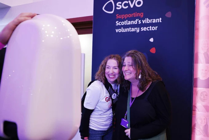 Two women: one on the left with curly red hair wears a white tshirt and one on the right with wavy brown hair is wearing a black top and lanyard. They stand close to each other and grin for a photo. They're standing in front of a banner that says "SCVO Supporting Scotland's vibrant voluntary sector"