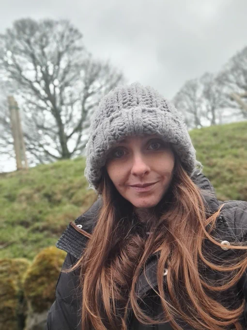A photo of me wearing a grey woolly hat. I have long brown hair and am standing with grass and trees behind me 