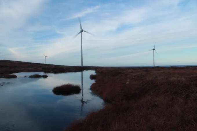Three thin wind turbines against a semi-cloudy sky, over peatland and bog, with a turbine reflected in the water.