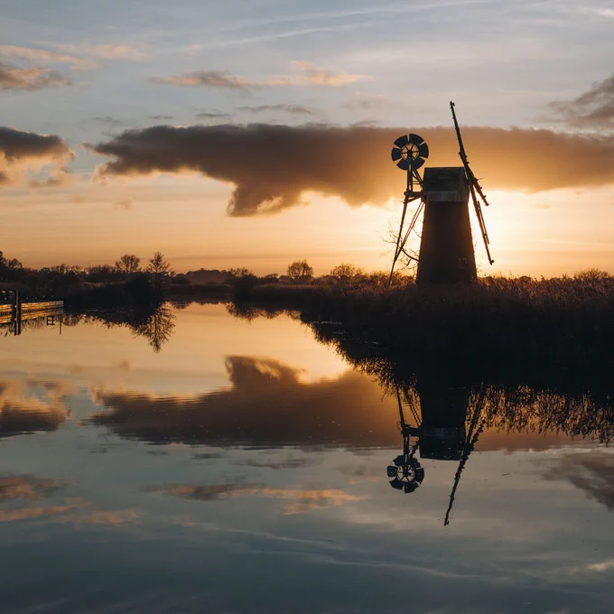 A serene view of a windmill at sunset, reflected in calm water. The sky is painted with soft oranges and yellows, while silhouetted reeds and trees frame the scene, evoking a tranquil countryside atmosphere.