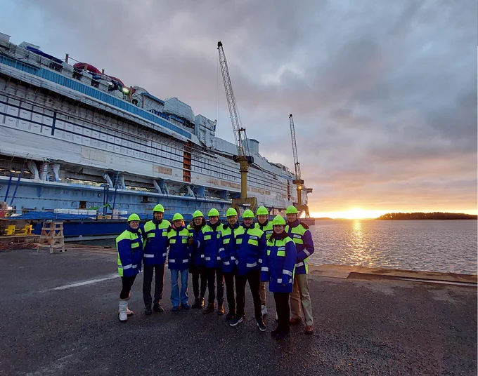 Kymmenen hengen porukka huomiovaatteissa Meyerin telakalla auringon laskiessa. Taustalla saaristoa ja rakenteilla oleva risteilyalus.