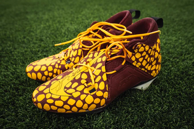 Photo of signed burgundy and gold Frankie Luvu cleats sitting on turf. 