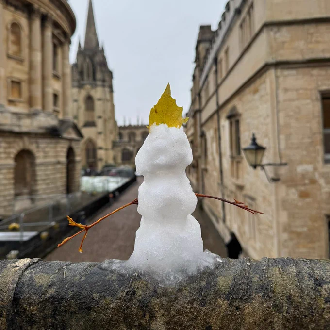 A small snowman decorated with twig arms and a yellow leaf as a crown, perched on a stone railing with Radcliffe Camera visible in the background on an overcast day.