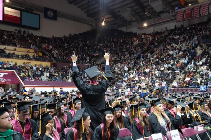 A graduating CALS student in cap and gown stands up and cheers toward the gathered audience