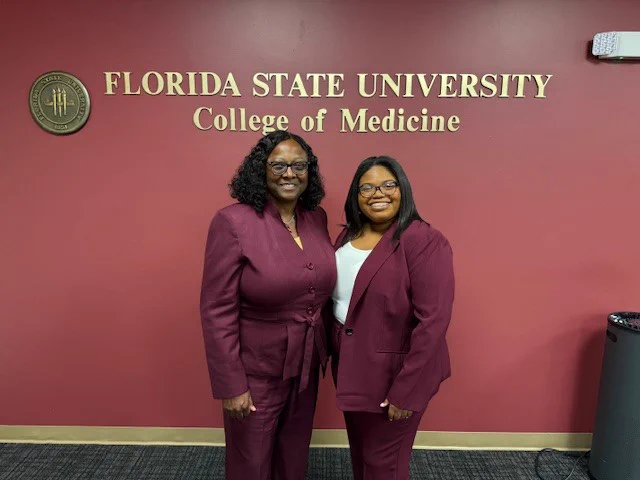 Two women in burgundy suits smiling in front of a red wall at the FSU College of Medicine.