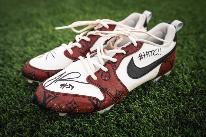 Photo of signed burgundy and white Jeremy Reaves cleats sitting on turf. 