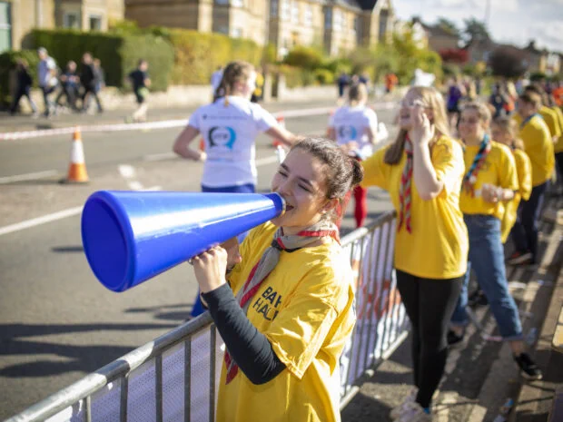A volunteer in a yellow t-shirt using a megaphone to cheer at the 2024 Bath Half, with other similarly dressed volunteers clapping by the roadside.
