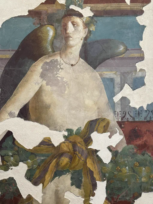 A winged female figure in part covered by a garland of vegetation with a knot of yellow and purple striped textile. Behind panels of blue and pink and red.