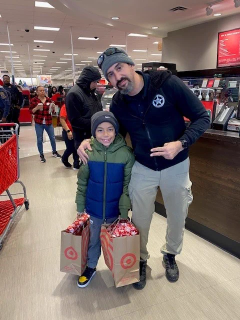 A Deputy U.S. Marshal stands with a child holding two bags with retail items. 