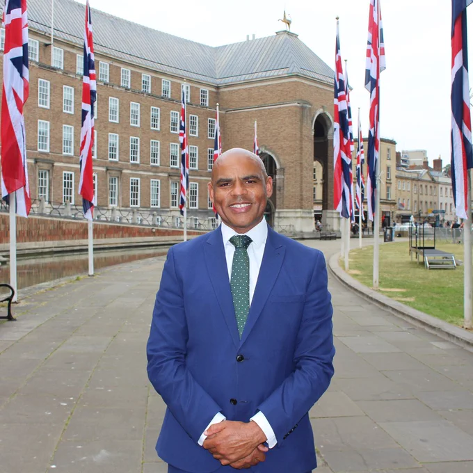 Marvin Rees OBE, the former Mayor of Bristol, is pictured, smiling, outside of City Hall with large Union Flags behind him.