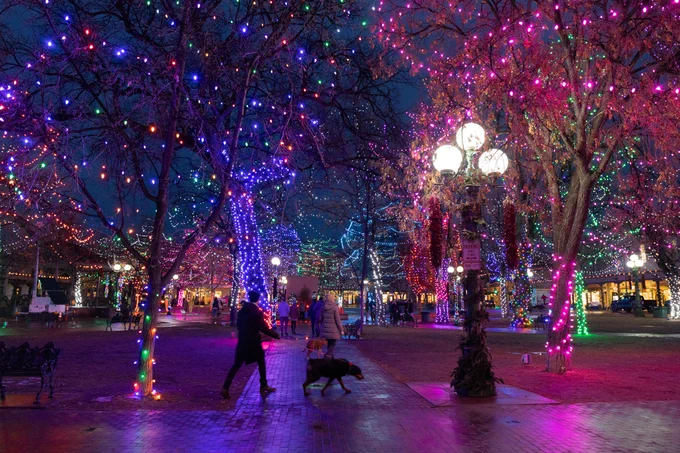 A man walks his dog across Santa Fe Plaza lit up with multi-colored holiday lights.