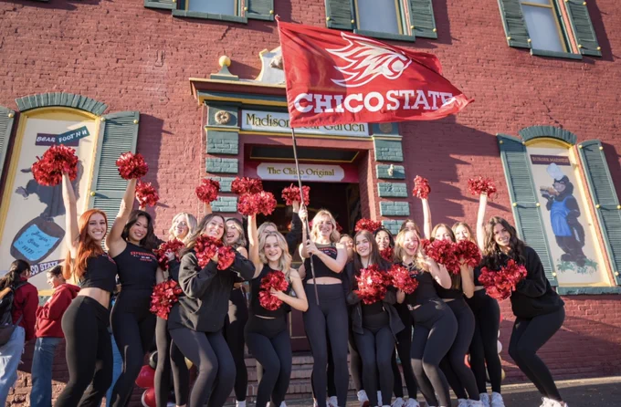 Chico State Expressions Dance Team stand in front of Madison Bear Garden waving a large Chico State flag and cheering at Chico State's Wildcat Tailgate.
