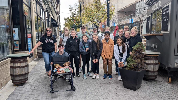 Group of teenagers smiling at camera. The wheelchair has creepy dolls sitting in it from a ghost tour