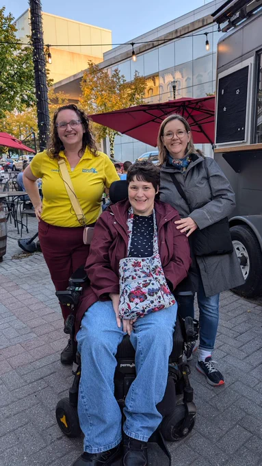 Woman in wheelchair smiling. Two women standing behind her smiling, one wearing yellow tour shirt.