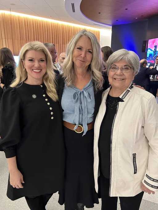 President Amy Parsons, Kim Jordan, and Polly Baca stand together indoors at an event, smiling for the photo. Attendees are visible in the background near a curved wood-paneled wall and a screen displaying a colorful graphic.