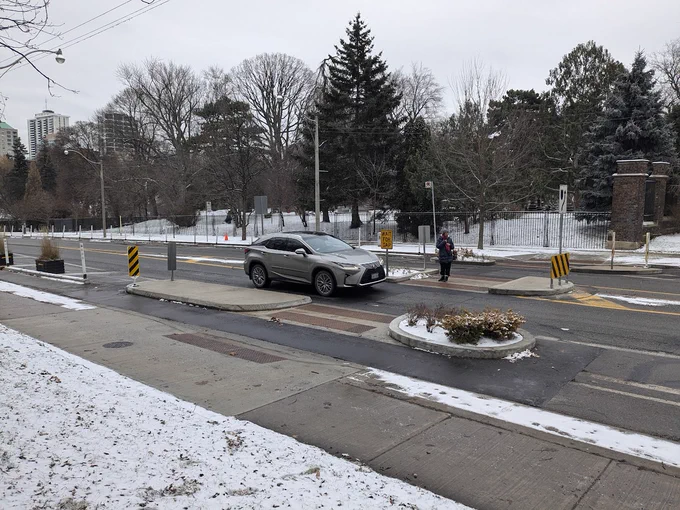 A person waits to cross a street at a pedestrian refuge island, which features tactile walking surface indicators, hazard marker signs, and planters. 