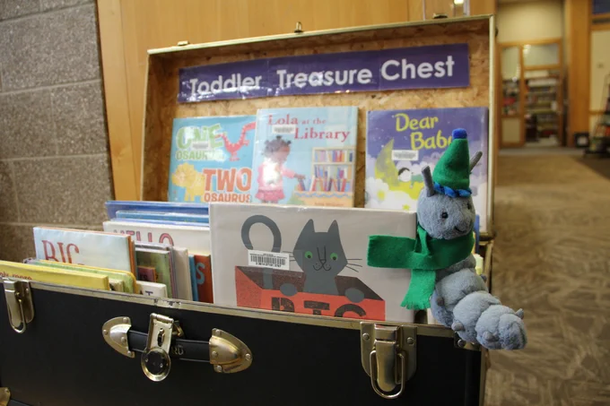 WCCLS mascot toy Wallace wearing a green hat and scarf on the Toddler Treasure Chest. You can see the over of some picturebooks in the Treasure Chest.