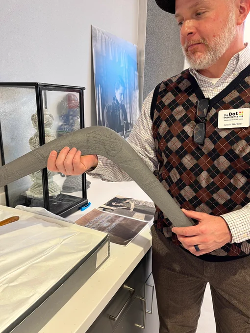A man with a Dot Experience nametag holds up a replica of a large boomerang as he stands near a display of historical artifacts.