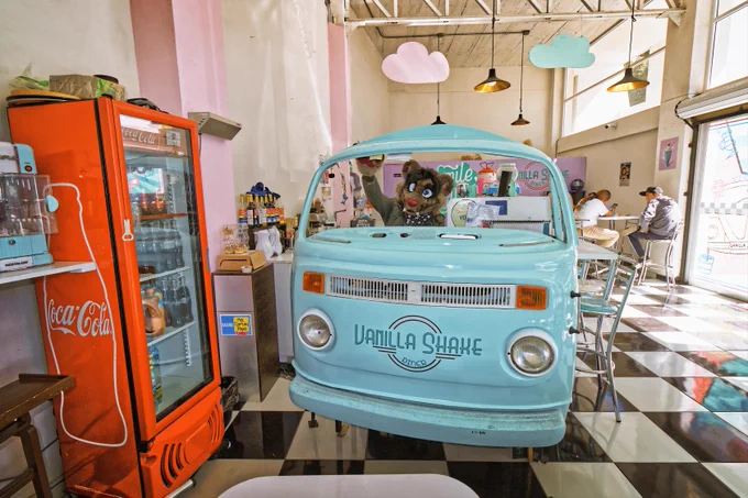 A friendly furry mascot welcomes customers from behind the counter of a charming retro diner, designed like a vintage Volkswagen bus.