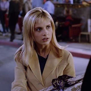 Buffy wears a tan coat and holds a wrapped gift — her hair is down and her bangs are so so so so tiny