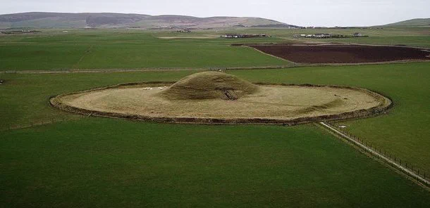 The chambered cairn of Maeshowe in Orkney. A circular site with a grass covered dome at the centre.