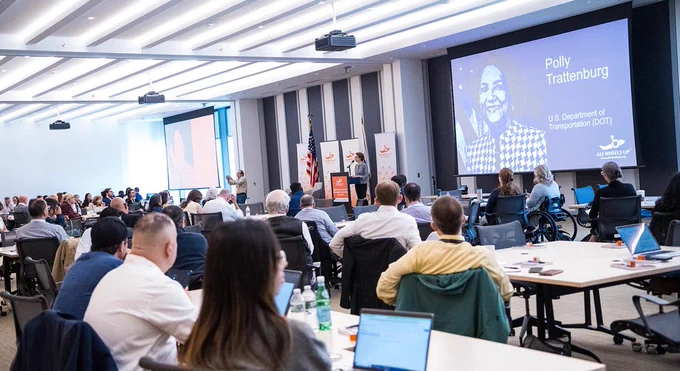 Image of our 2024 Global Forum Attendees listening to Polly Trottenburg give her speech with her name written on the screen behind her.