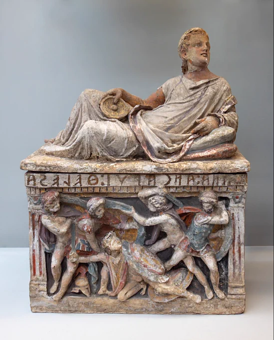 Absolutely gorgeous painted terracotta cinerary urn with the figure of a young fair-haired, blue-eyed man reclining on the lid - propped up by two pillows - and a molded panel on the chest. The figure wears a white tunic (chiton) with two purple clavi (vertical stripes), a mantle - possibly a toga - with a purple border and a gold finger-ring, and holds a libation bowl (phiale). The name of a woman Thana Ancarui Thelesa is painted on the chest of the urn and a scene of five colorful embattled warriors appears below. How did the figure of a man on the top get mixed with the chest bearing a woman's name?

Etruscan, about 150-100 BCE, from Chiusi, Italy. 

British Museum (1926,0324.124)