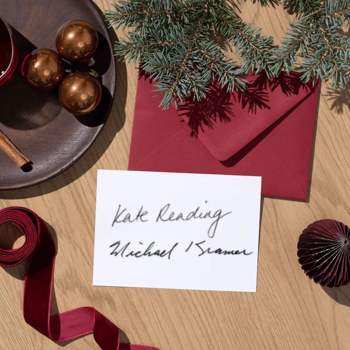 Festive gold, red, and green baubles on a wood table, including small gold ornaments, a pine bough, and a red envelope. A white sheet of paper is on top of the red envelope. The white sheet of paper has the signatures of Kate Reading and Michael Kramer.