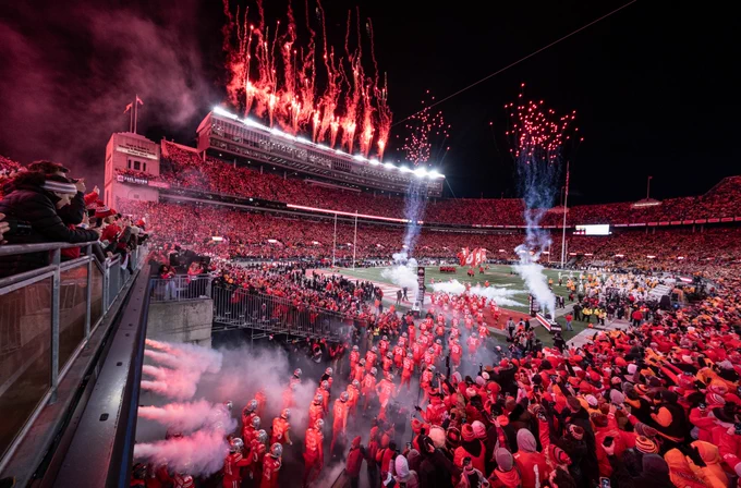 Ohio Stadium filled with fans wearing Scarlet. The football team is running out onto the field and pyrotechnics are going off.