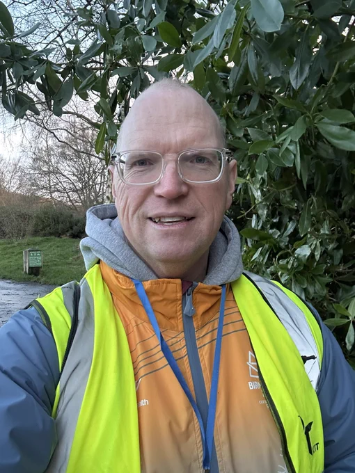 Photograph shows the author smiling outside with a tree behind. The author is wearing a grey hoodie with a Birmingham 2022 Commonwealth Games volunteer jacket and a high visibility vest and lanyard with blue string.