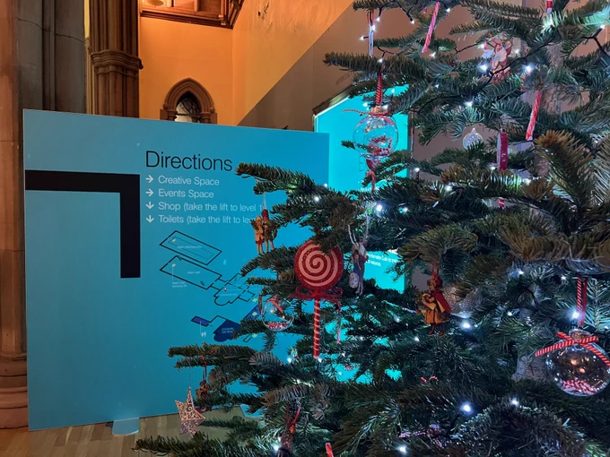 A Christmas tree in the Hunterian Museum, decorated with red and white candy canes and baubles. Beside the tree, a blue text panel is displaying directional signage to the Museum. 