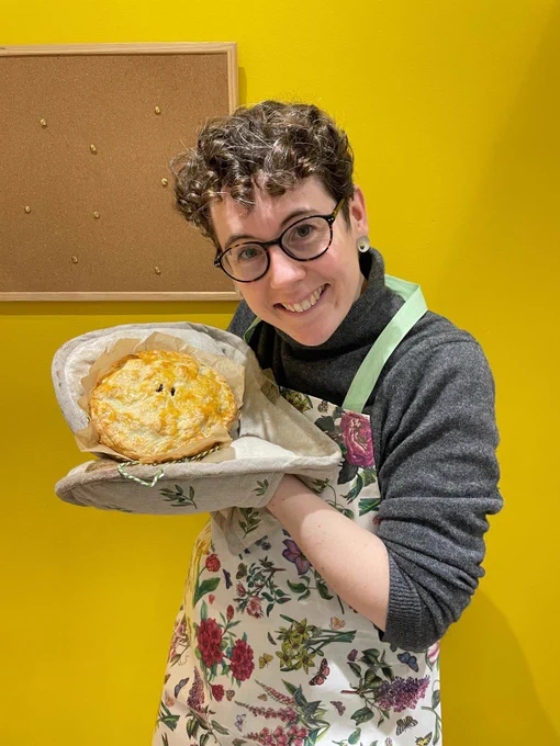 A smiling white woman with short curly hair in a flowery apron, showing off a large pie