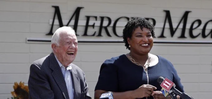 Jimmy Carter laughing and Stacey Abrams smiling in front of a new medical clinic for the uninsured.