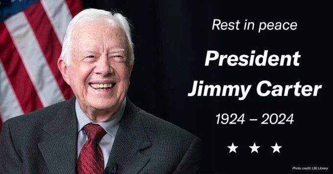 Image of a smiling President Jimmy Carter in a suit, with an American flag in the background and text that reads "Rest in peace President Jimmy Carter 1924 – 2024." Three stars are at the bottom. Photo credit to LBJ Library.