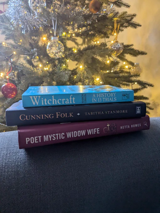 A pile of three books on the arm of a sofa, in front of a Christmas Tree. 'Witchcraft: A History in 13 Trials by Marion Gibson', 'Cunning Folk' by Tabitha Stanmore and 'Poet Mystic Widow Wife' by Hetta Howes