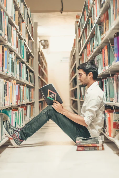 A man sitting on the floor of a library, between two aisles of books. He is reading a book, and a wearing glasses, a white shirt, blue jeans and trainers. Photo by Dollar Gill on Unsplash. 